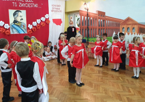 Dziewczynki w czerwonych sukienkach oraz chłopcy ubrani na galowo wykonują taniec "Mazur".